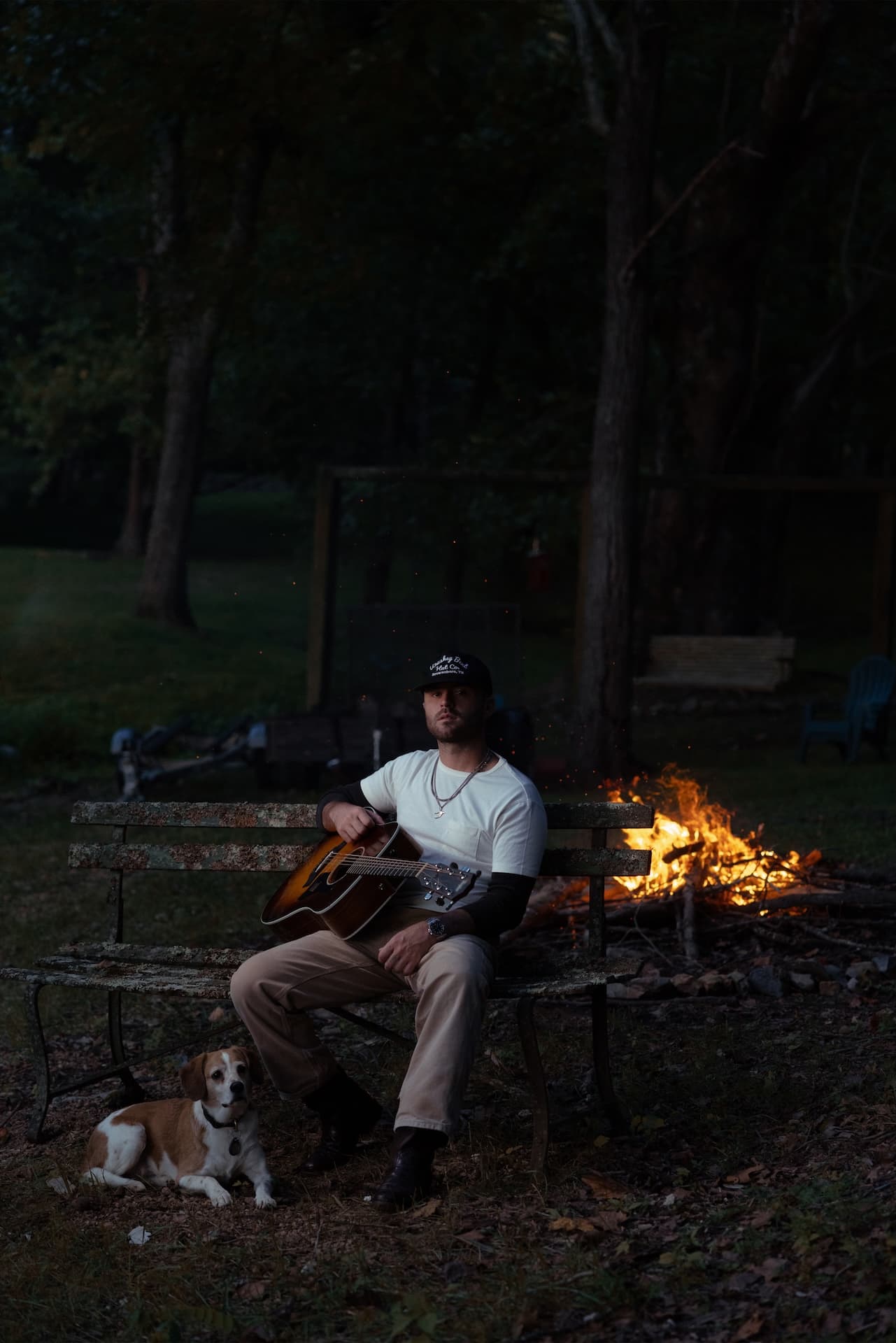 Greylan James with his guitar on a bench beside a bonfire at dusk, a beagle at his feet