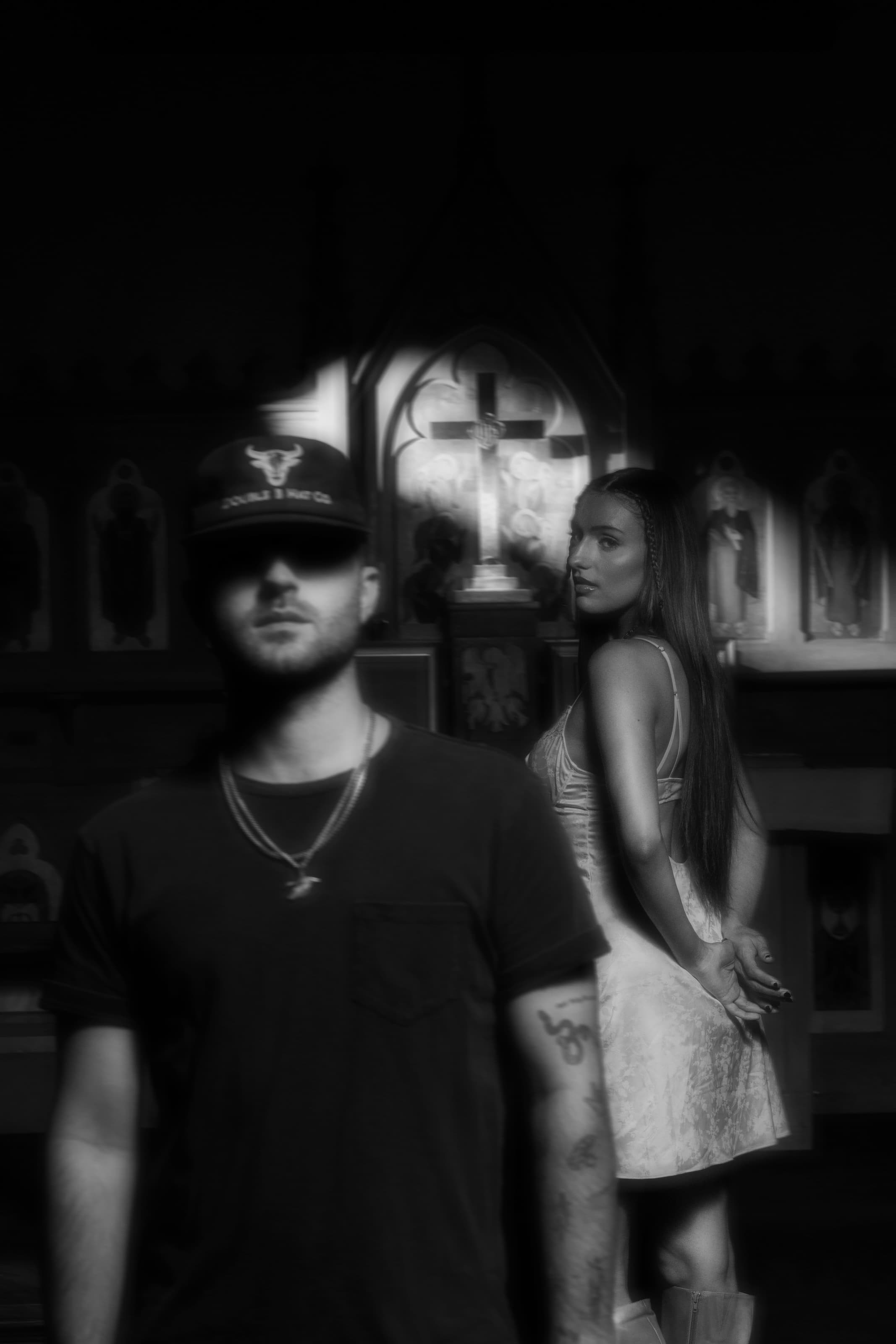 Greylan James in a black ball cap and t-shirt, with Gabriella Rose in a white slip dress looking back over her shoulder, inside a candlelit chapel